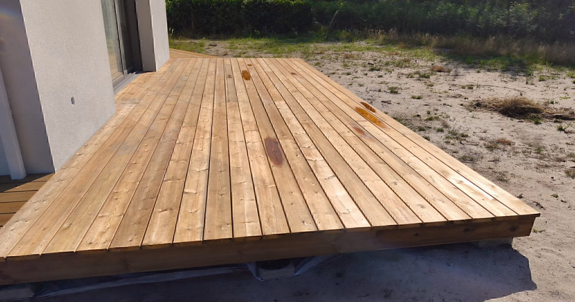 Grande terrasse en bois clair longeant la façade blanche d'une maison avec une baie vitrée.