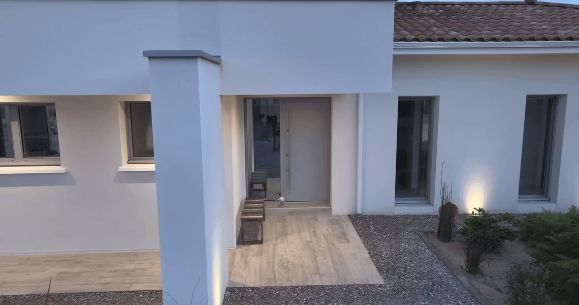 Façade blanche moderne avec porte d'entrée grise, fenêtres à encadrements et parvis en bois.