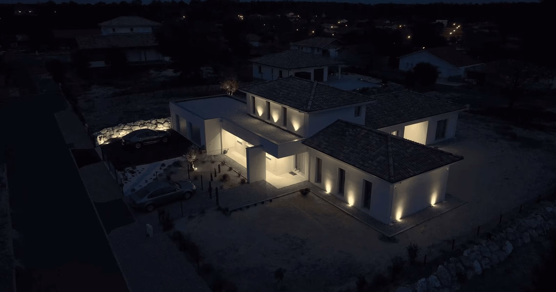 Vue aérienne de nuit de la villa moderne éclairée avec des spots en façade et des voitures garées.