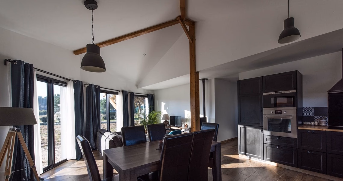 Grand salon lumineux avec charpente apparente, table à manger en bois et cuisine ouverte noire équipée.