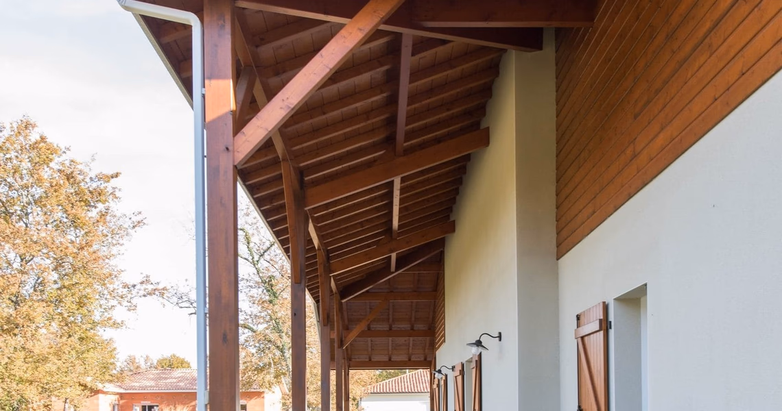 Grande charpente en bois apparente protégeant la terrasse d'une maison aux murs blancs et bardage.