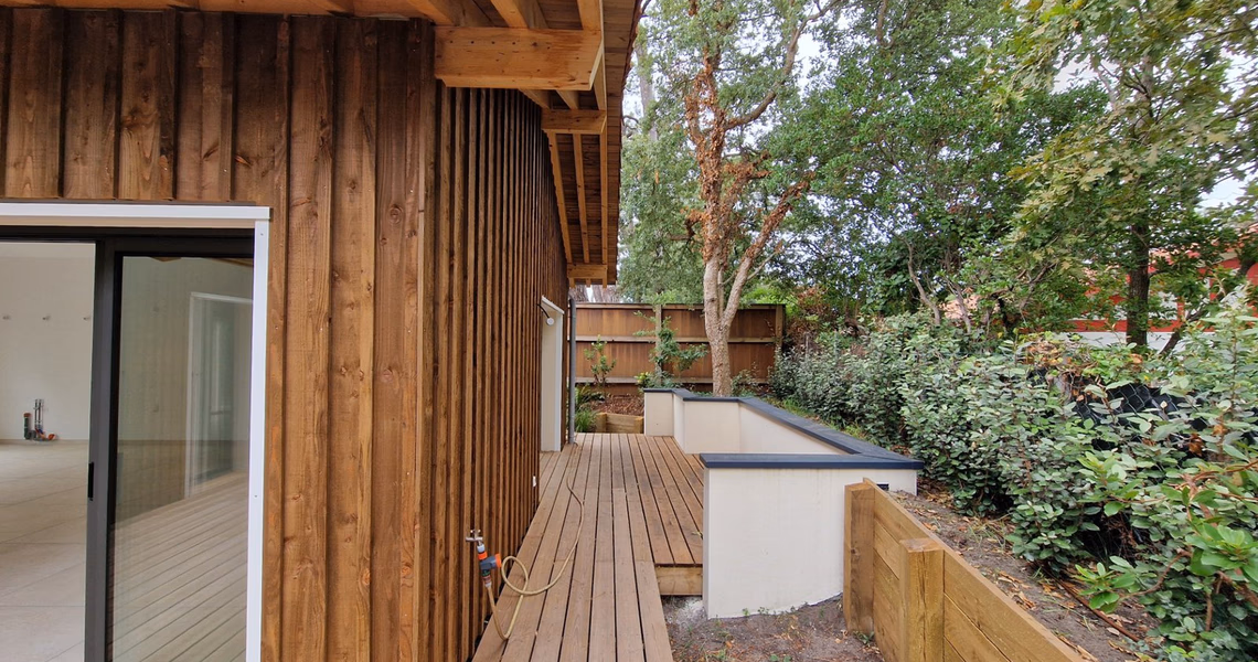 Façade en bois clair avec une large baie vitrée et une terrasse en bois bordée de verdure.