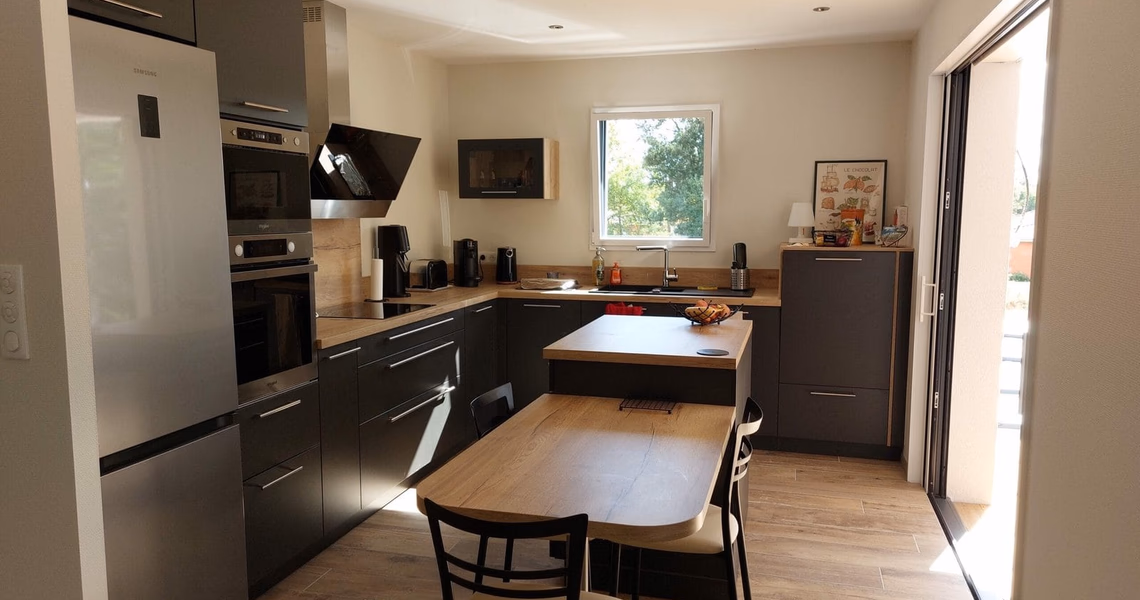 Cuisine moderne en L avec meubles sombres, plan de travail en bois et table à manger intégrée.