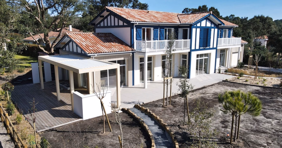 Grande villa blanche à étage avec colombages bleus, toiture en tuiles et terrasse en bois.
