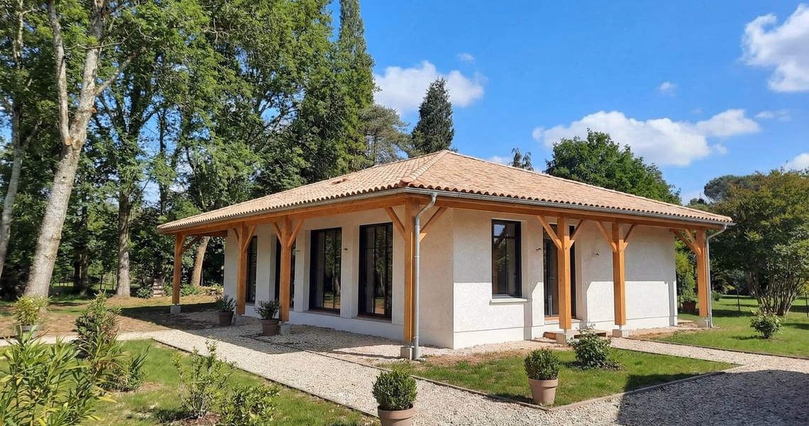 Maison de plain-pied aux façades claires avec toiture en tuiles et porche en bois soutenu par des piliers.