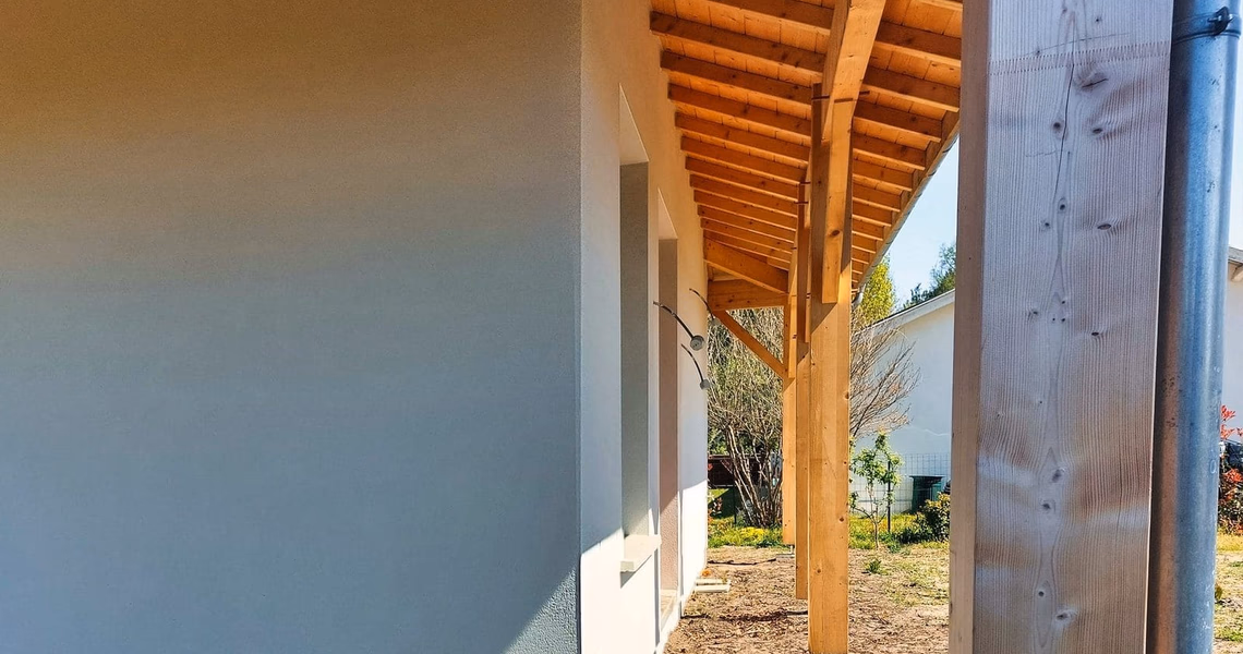 Façade blanche d'une maison avec une avancée de toit en bois soutenue par des poutres.