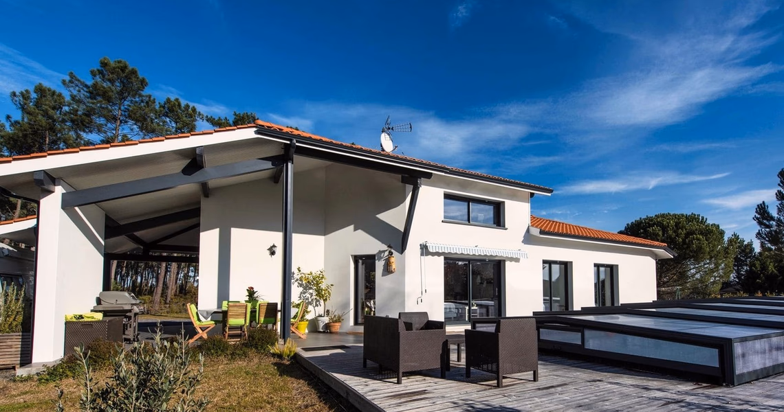 Façade blanche d'une maison contemporaine avec terrasse en bois, mobilier de jardin et piscine couverte.