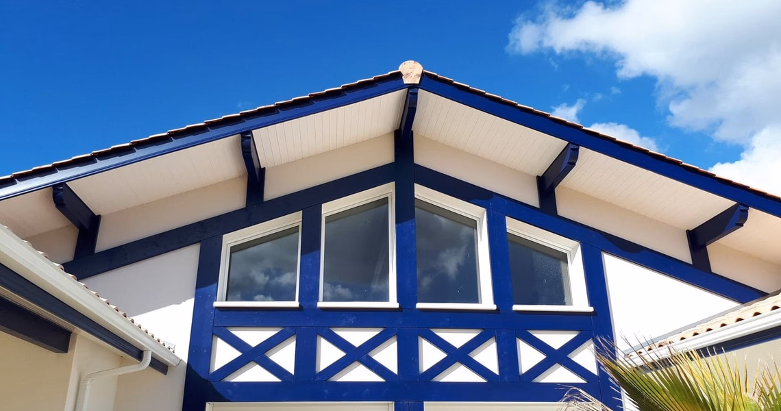 Gros plan sur la charpente apparente bleu foncé, les boiseries blanches et les fenêtres de la villa.