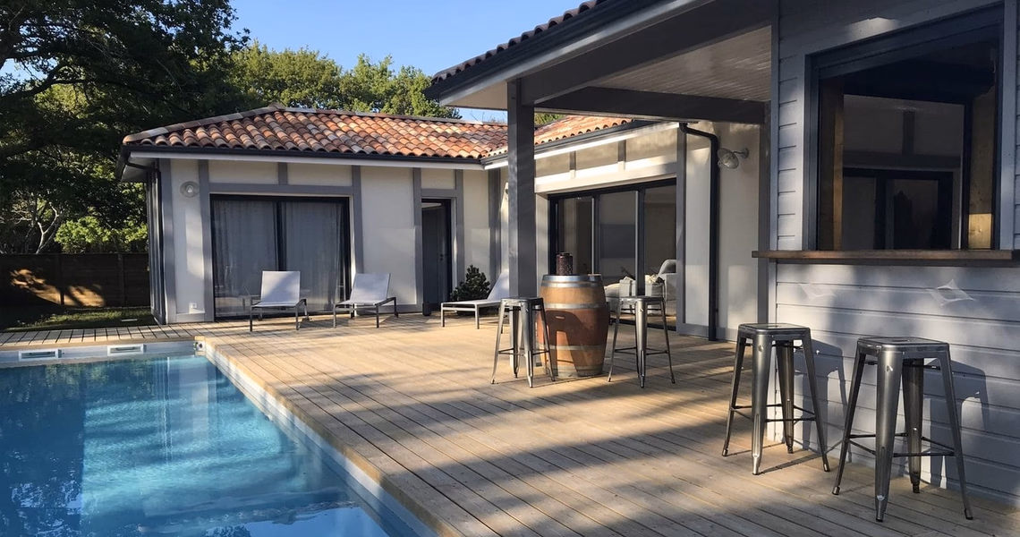 Maison moderne en L avec terrasse en bois, piscine et bar extérieur équipé de tabourets métalliques.