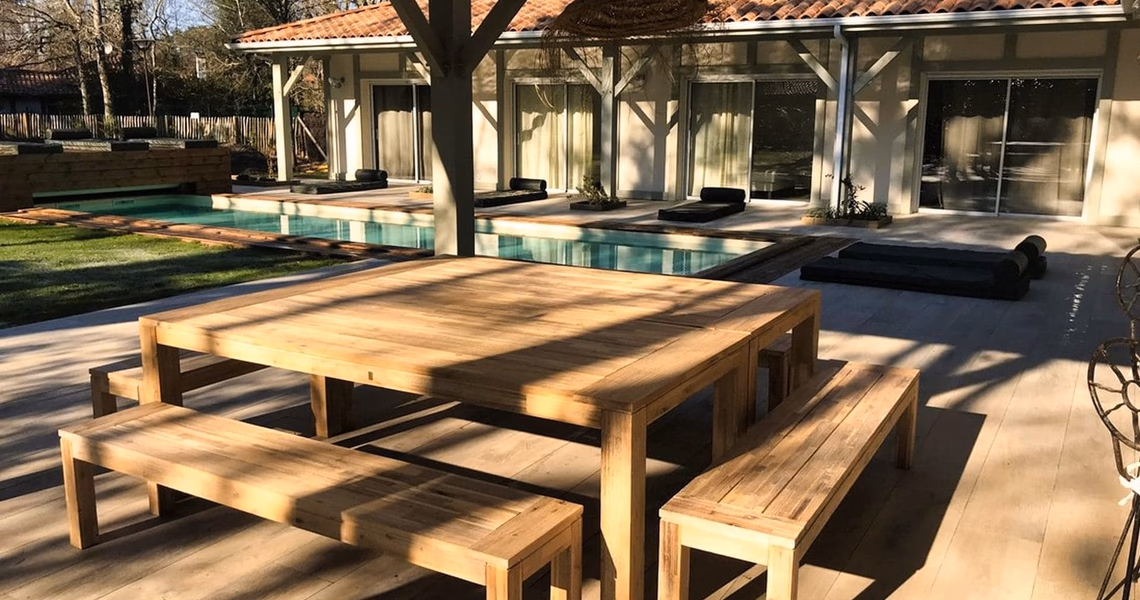 Grande table en bois sur une terrasse carrelée avec piscine et façade de maison de plain-pied.