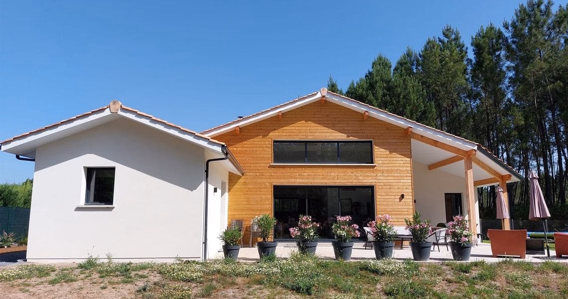 Façade de maison de plain-pied avec bardage bois, enduit blanc, grande baie vitrée et terrasse fleurie.