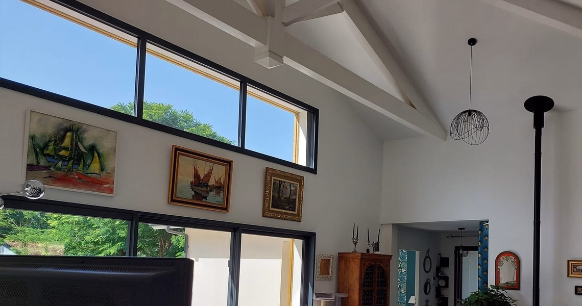 Salon lumineux avec grande hauteur sous plafond, charpente apparente, larges baies vitrées et poêle.