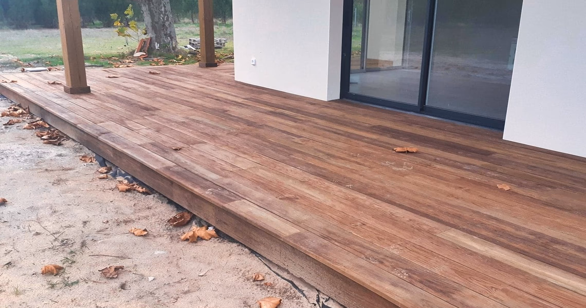 Grande terrasse en bois avec poteaux de soutien le long d'une façade blanche et d'une baie vitrée.