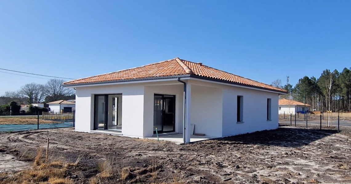 Maison de plain-pied aux murs blancs et toiture en tuiles orange avec menuiseries noires et terrain brut.