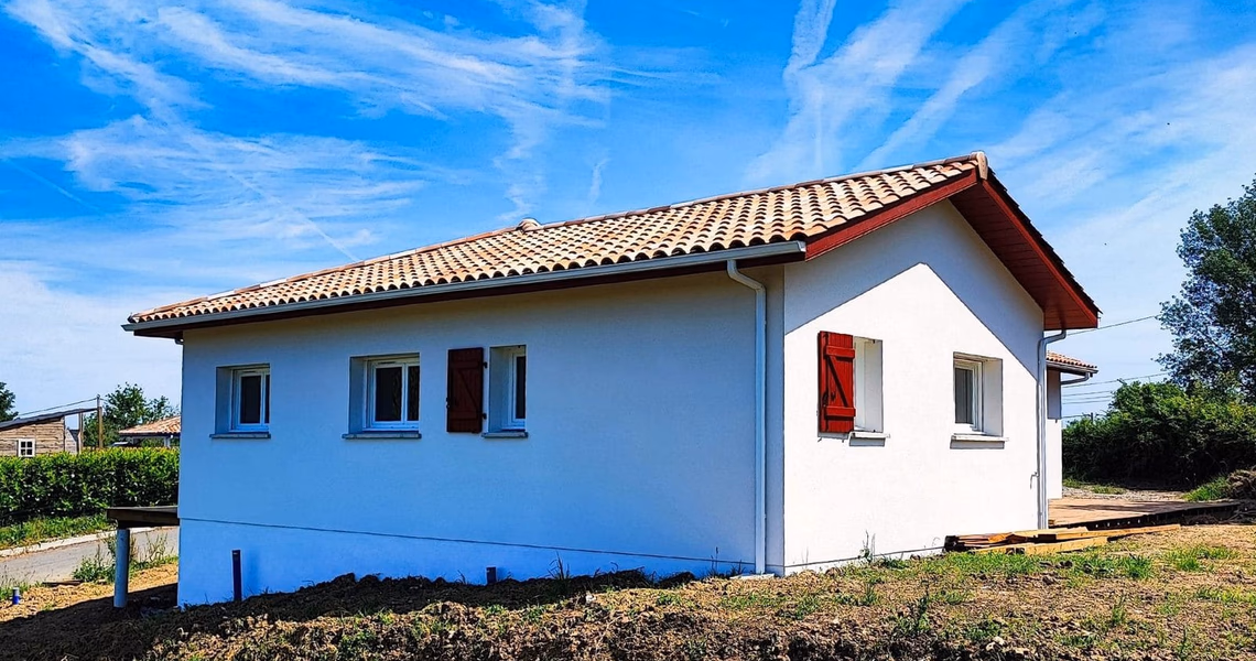 Façade latérale d'une maison individuelle blanche aux volets rouges sous un ciel bleu dégagé.