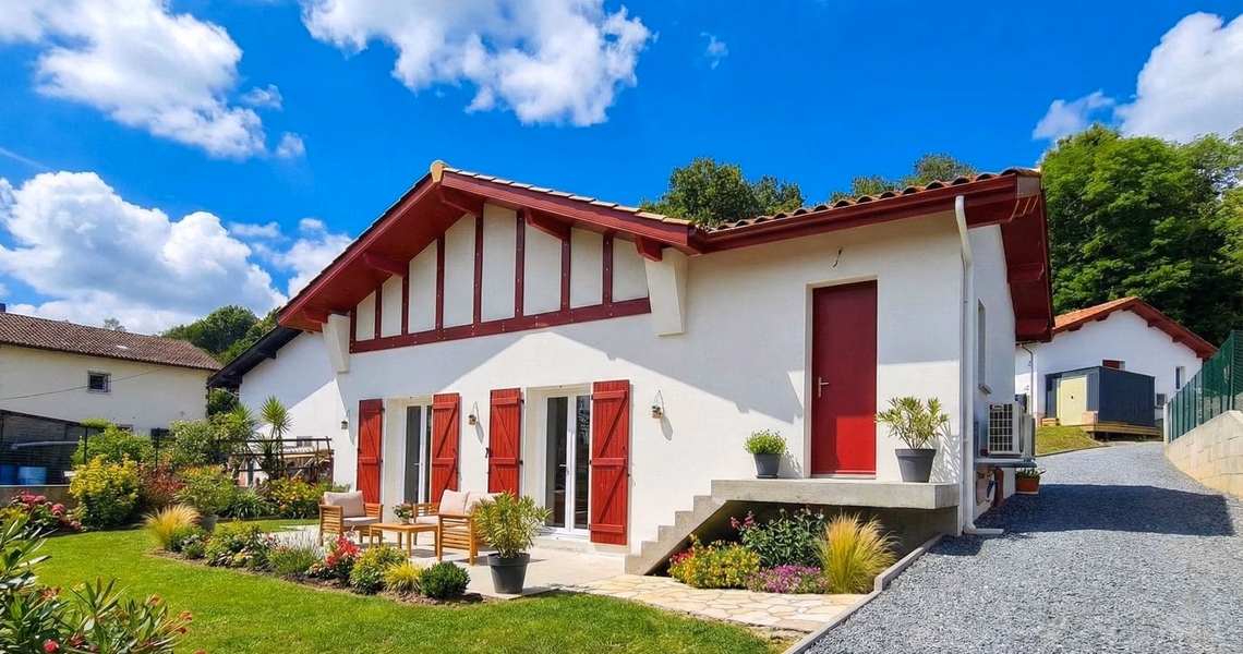 Façade de maison blanche et rouge style basque avec porte rouge, volets et salon de jardin.
