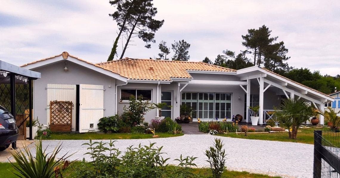 Maison de plain-pied grise à toit en tuiles, avec grande terrasse couverte et jardin paysager.