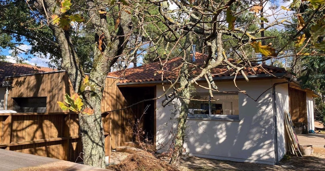 Façade de plain-pied en bois et enduit clair avec toiture en tuiles cachée par des arbres.