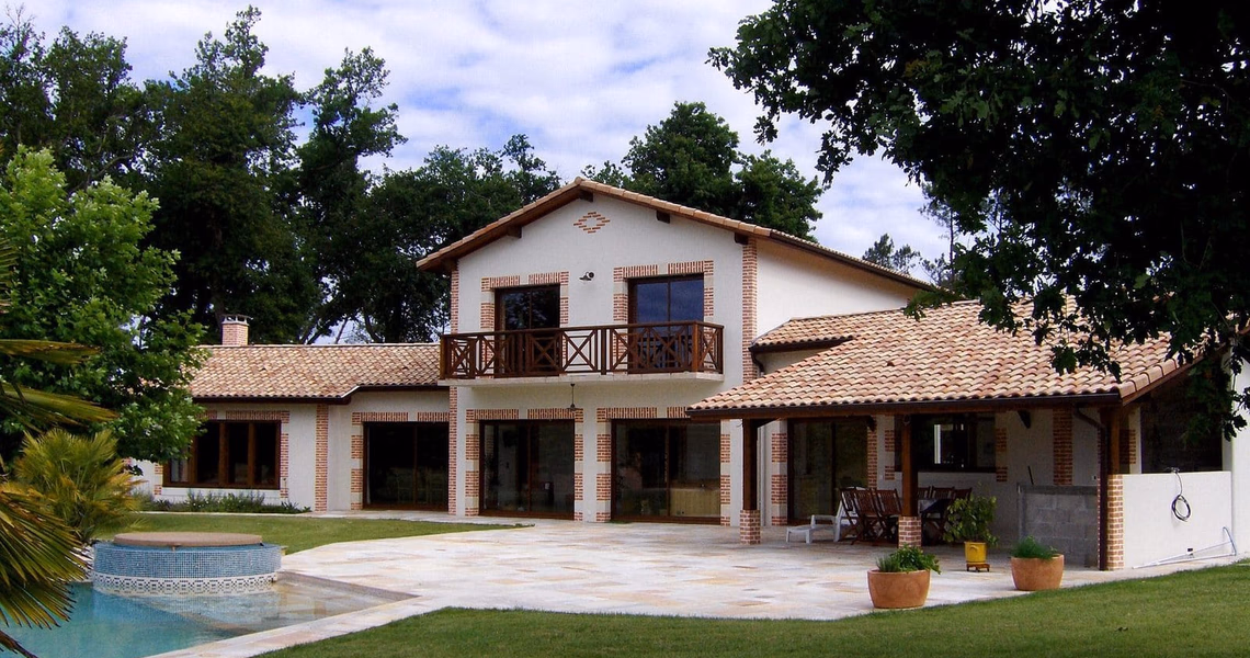 Grande maison landaise blanche à étage avec toit en tuiles, terrasse dallée et piscine au premier plan.