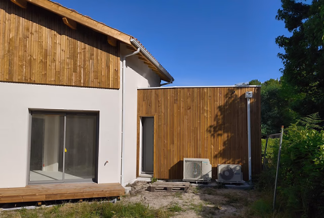 Façade d'une maison à étage avec bardage bois vertical, enduit blanc et deux unités extérieures de pompe à chaleur.