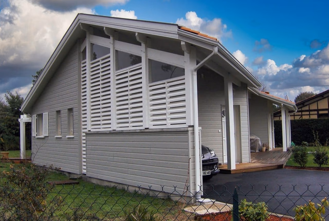 Maison individuelle à ossature bois grise dotée de larges baies vitrées et d'un carport latéral.