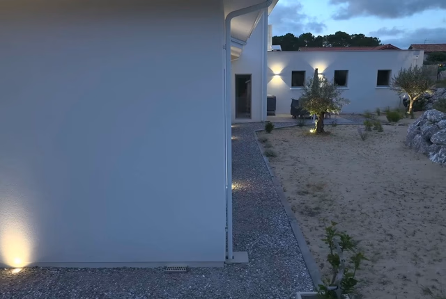 Façade blanche éclairée d'une villa contemporaine avec allée de graviers et jardin paysager au crepuscule.