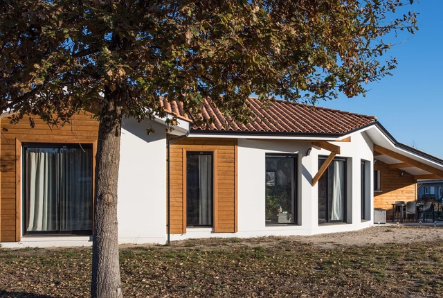 Façade de maison de plain-pied avec enduit blanc, habillages en bois et terrasse couverte.