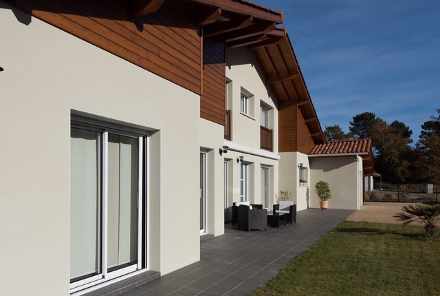 Façade de la maison à étage avec enduit clair, bardage bois sous toiture et grande terrasse carrelée.