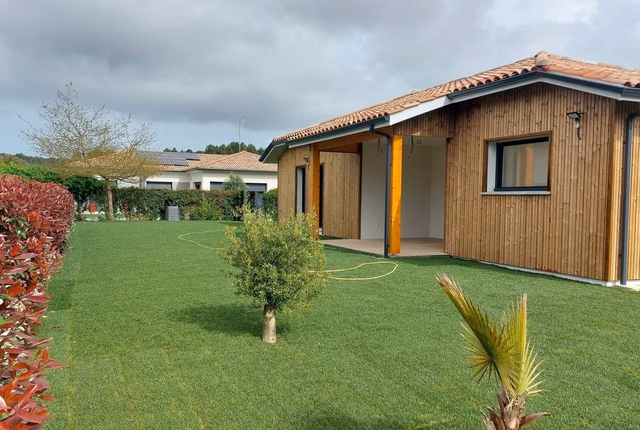 Maison de plain-pied à bardage bois vertical avec toiture en tuiles, terrasse couverte et jardin.