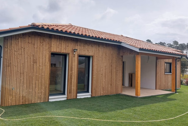 Façade latérale d'une maison avec bardage bois vertical, grandes baies vitrées et toiture en tuiles.