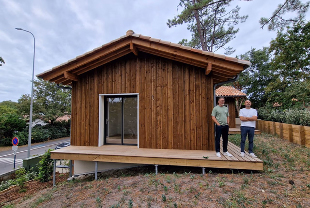 Façade en bois avec une baie vitrée ouvrant sur une terrasse en bois surélevée par des piliers.