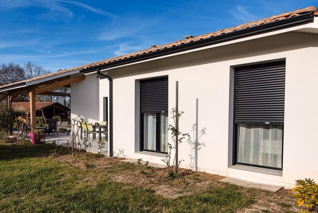 Façade blanche d'une maison de plain-pied avec deux fenêtres à volets roulants et terrasse couverte.