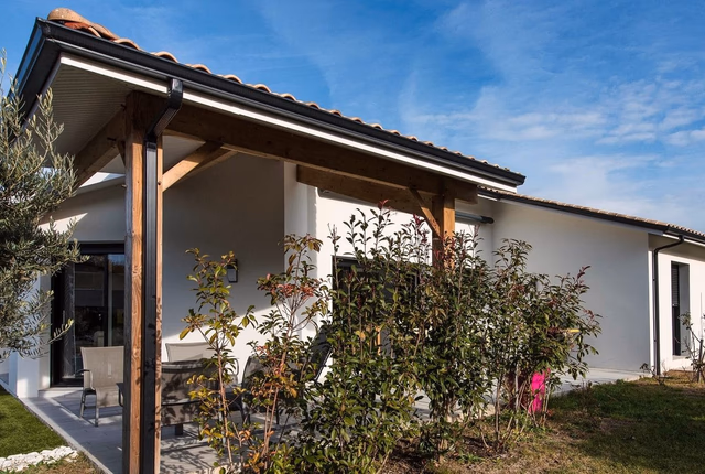 Façade de maison de plain-pied blanche avec porche en bois et terrasse carrelée devant un jardin.