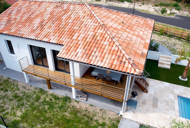 Vue aérienne d'une maison blanche avec toit en tuiles, terrasse en bois et piscine extérieure.