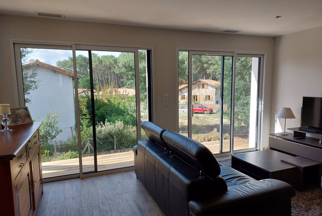 Salon lumineux avec un grand canapé noir, ouvert sur une terrasse en bois et un paysage arboré.