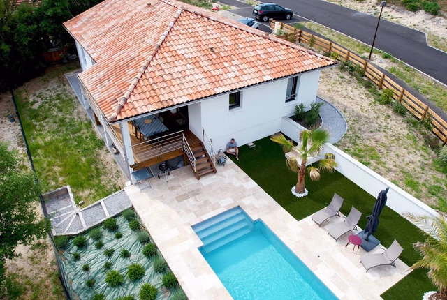 Vue aérienne d'une maison blanche à toit en tuiles avec piscine, terrasse dallée et transats.