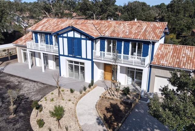 Grande villa blanche à étage avec toiture en tuiles romanes, volets bleus et terrasses à balustrades.