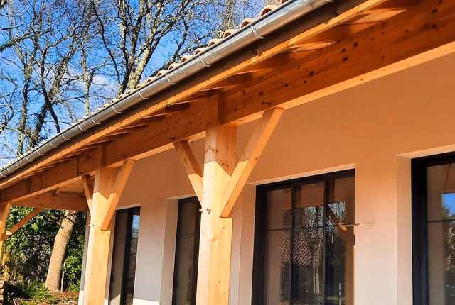 Façade blanche d'une maison de plain-pied avec auvent en bois, poteaux massifs et grandes baies vitrées.