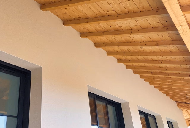 Avancée de toit en bois brut avec poutres apparentes surplombant la façade blanche de la maison.