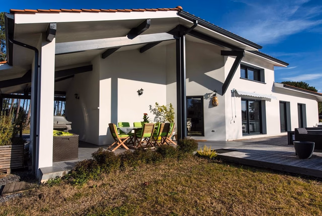 Façade blanche d'une maison moderne avec grande terrasse en bois, mobilier de jardin et pergola.
