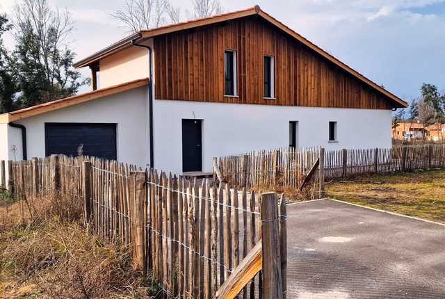 Villa R+1 moderne aux murs blancs et bardage bois en pignon, entourée d'une clôture en bois.
