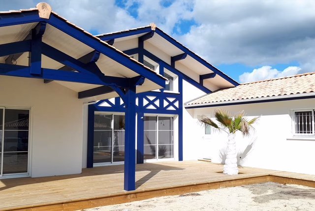 Grande terrasse en bois devant une villa blanche aux boiseries bleues et toiture en tuiles.