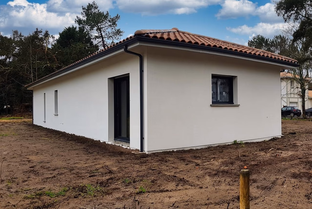 Façade latérale d'une maison de plain-pied en crépis blanc avec toit en tuiles et terrain en terre.
