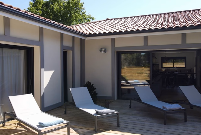 Terrasse en bois avec quatre bains de soleil devant la façade enduite blanche et grise.