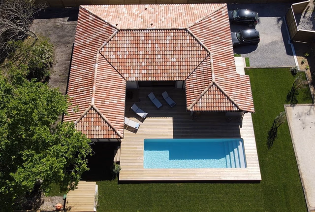 Vue aérienne d'une maison moderne en L avec toiture en tuiles, terrasse en bois et piscine rectangulaire.