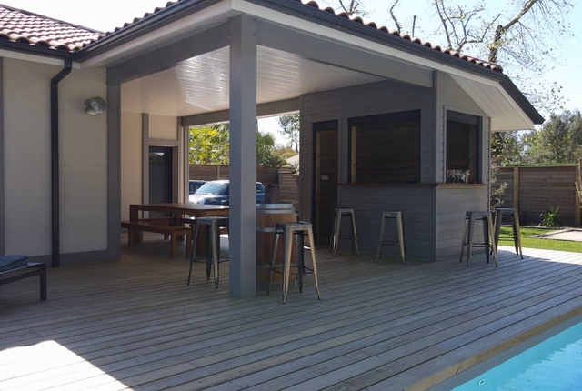 Grande terrasse en bois avec piscine et bar extérieur couvert par une charpente grise.