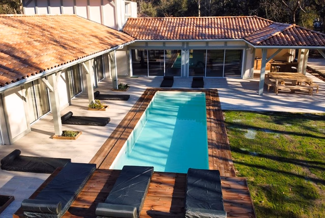 Grande maison en U avec toiture en tuiles rouges, piscine rectangulaire et terrasse en bois.
