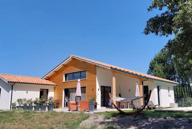 Maison de plain-pied en bois et enduit blanc avec toit en tuiles, terrasse et jardin arboré.