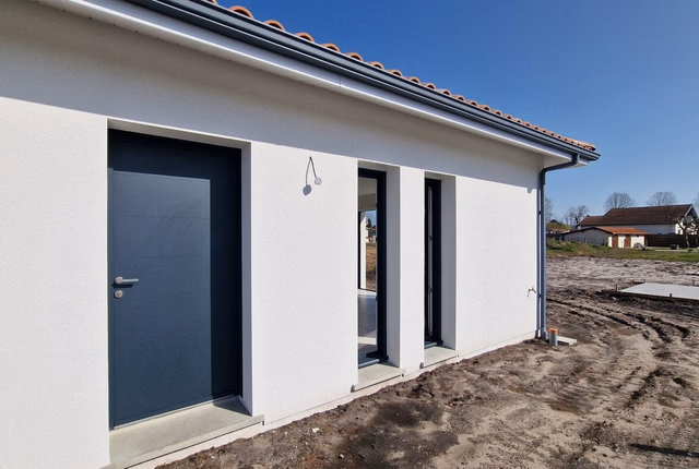 Façade de maison de plain-pied blanche avec porte d'entrée et menuiseries grises, et terrain en terre.