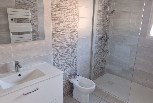 Salle de bain carrelée avec meuble vasque blanc, douche à l'italienne vitrée et bidet.